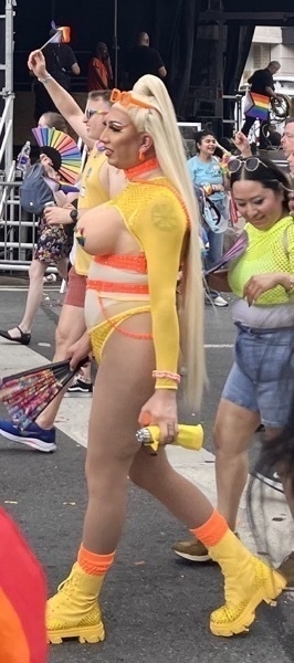 A trans woman with long blond pony tail, dramatic eye liner, and bright yellow combat boots. She wears a fish net bikini, with breasts exposed, nipples covered with rainbow paint. She walks confidently. 