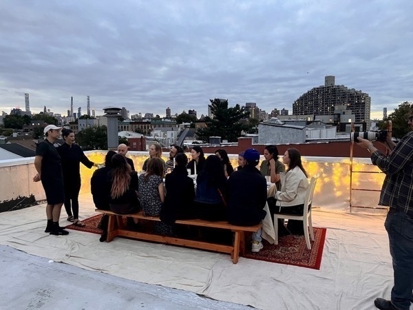 A group of people seated around a table on a rooftop under a cloudy sky, with one person standing and speaking. String lights decorate the scene, and city buildings are visible in the background.