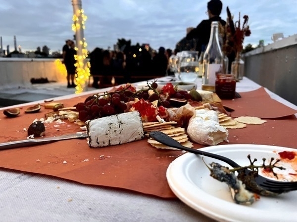 A table holds a partially eaten cheese and crackers platter, surrounded by wine glasses and bottles. Guests stand in the background under a cloudy sky, with lights and flowers providing decoration.