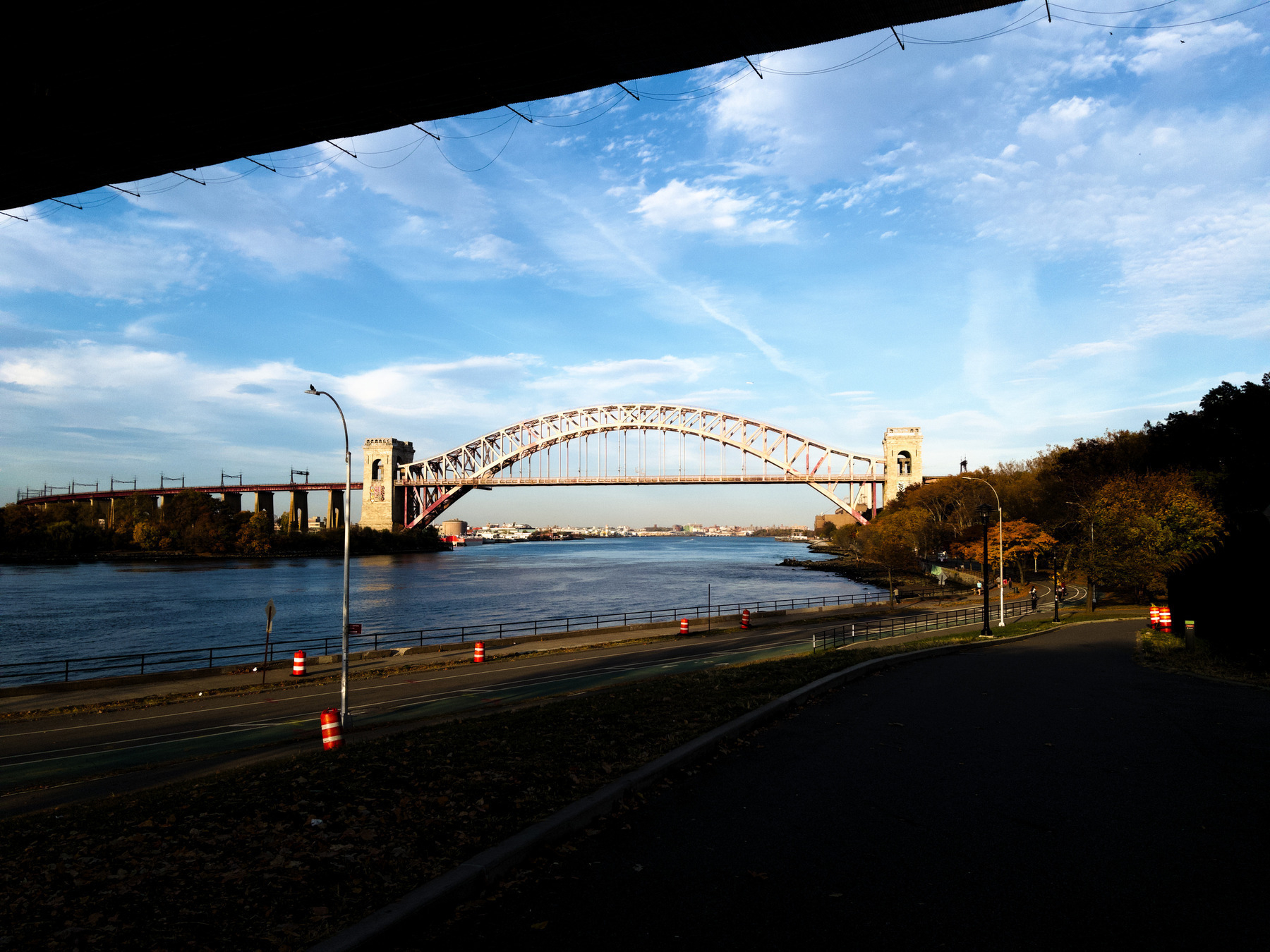 Auto-generated description: A large arch bridge spans a wide river under a cloudy blue sky, with trees and a road in the foreground.