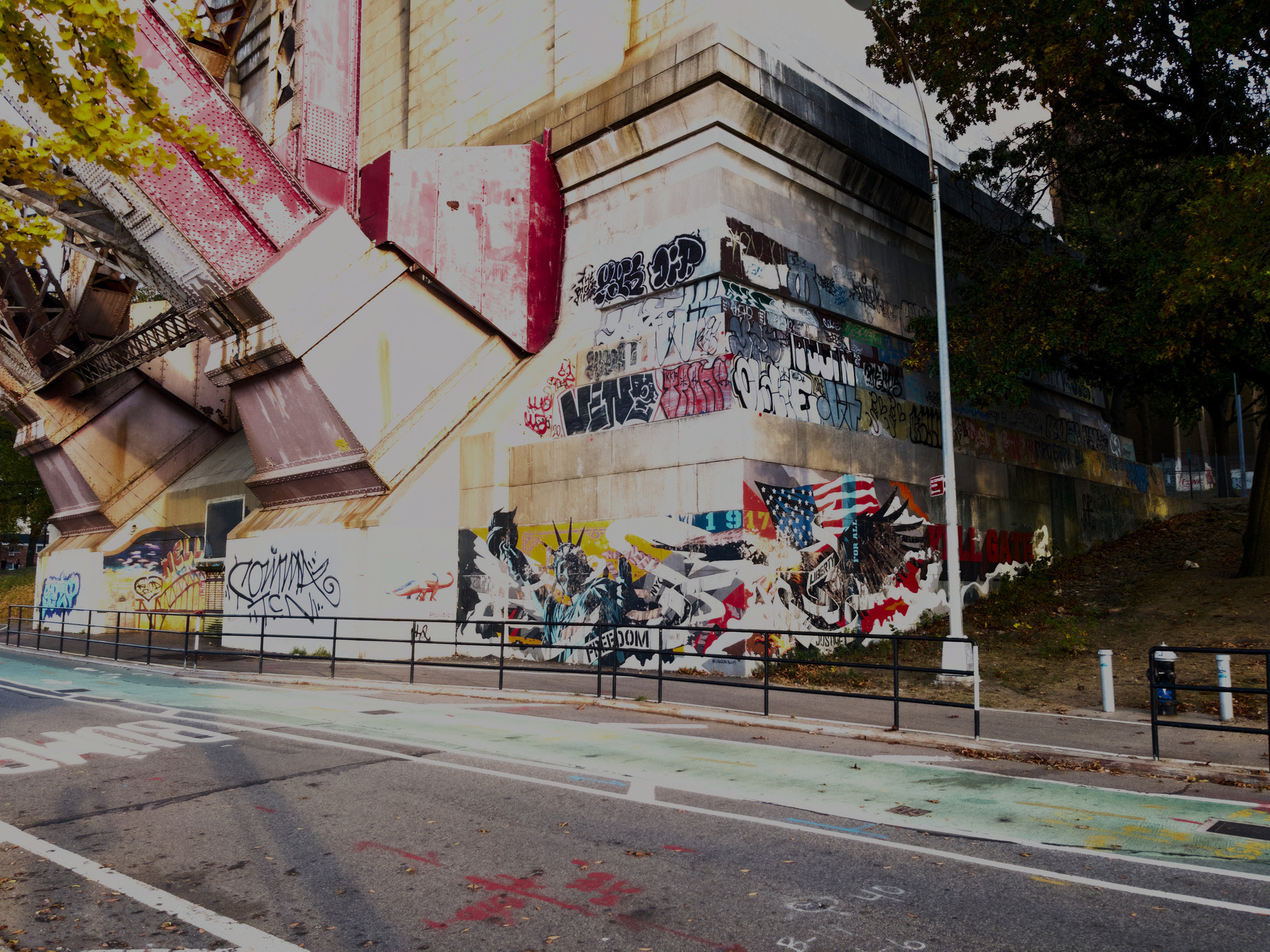 Auto-generated description: A graffiti-covered bridge support structure is surrounded by trees and bordered by a road with bike lanes.