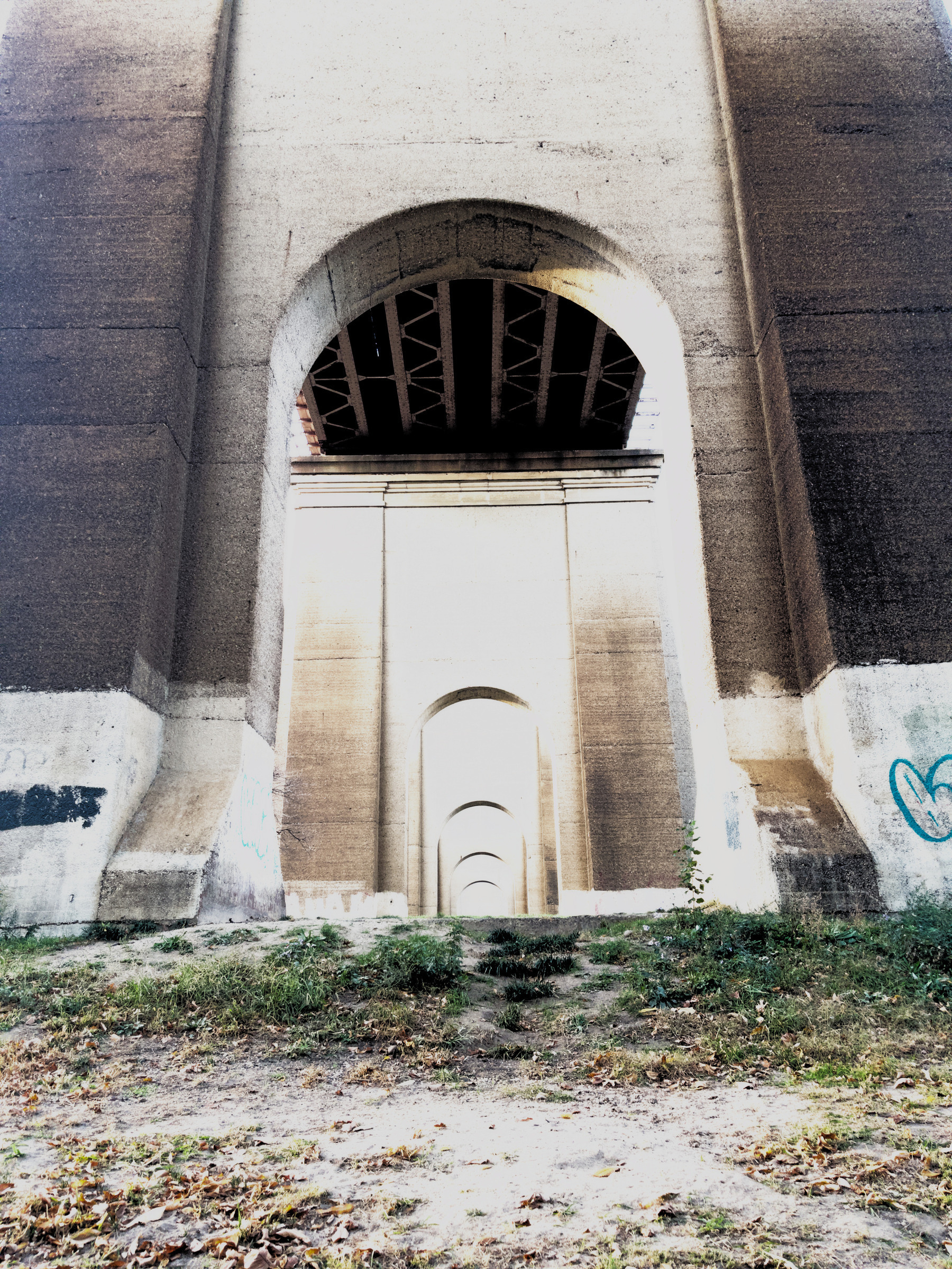 Auto-generated description: A series of large concrete arches creates a tunnel-like effect beneath a bridge, with graffiti and plants visible on the ground.