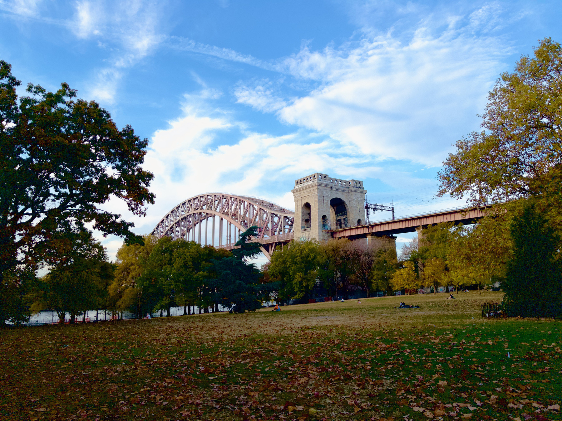 Auto-generated description: A large arch bridge spans across a park area with trees and a grassy field, set against a partly cloudy sky.