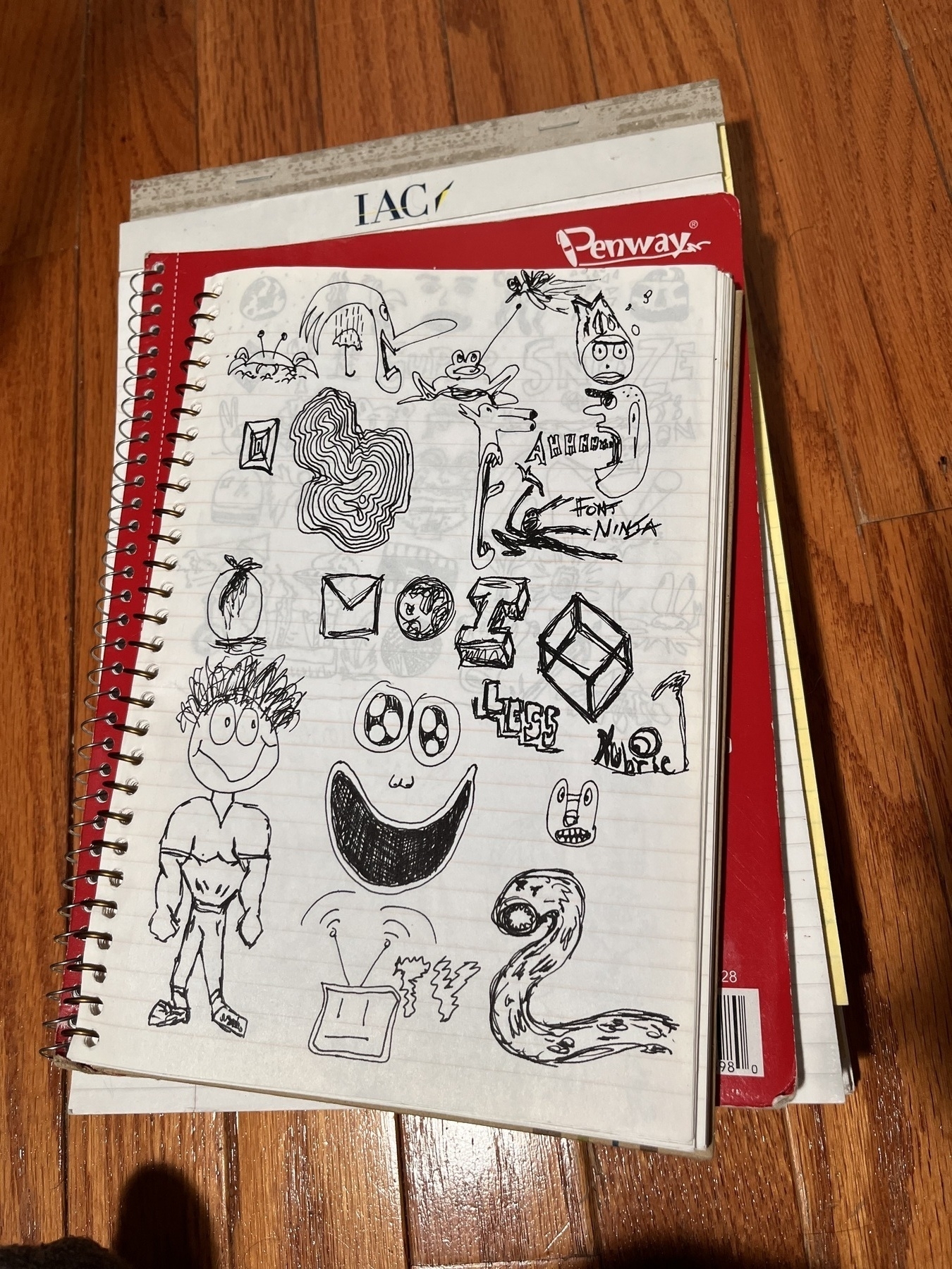 Auto-generated description: A spiral notebook page filled with various black ink doodles, including abstract shapes, faces, and text, rests on a wooden floor with other notebooks underneath.