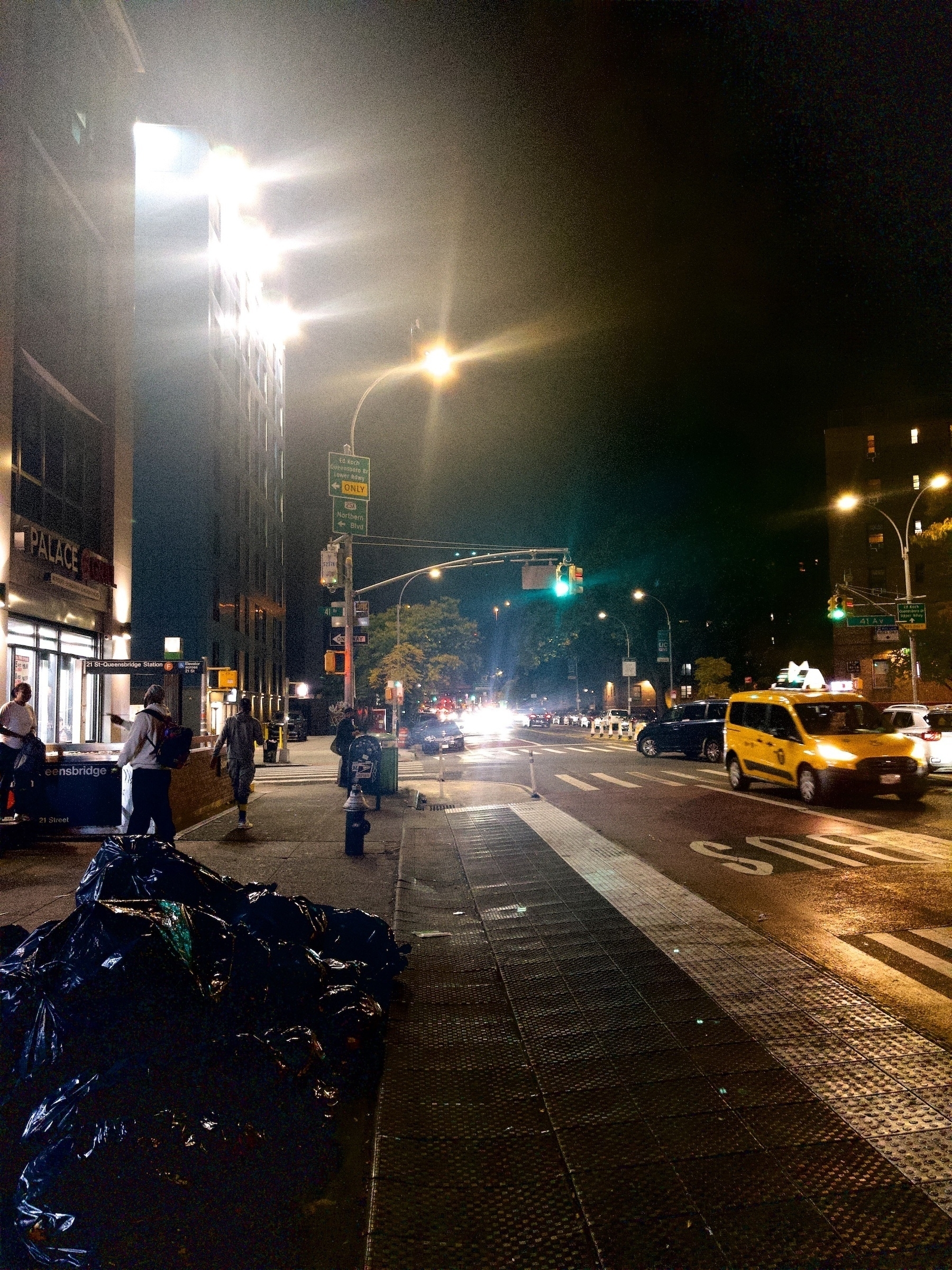 Auto-generated description: A nighttime urban street scene shows a bustling city street with cars, including a yellow taxi, and pedestrians under bright streetlights, alongside bags of trash on the sidewalk.