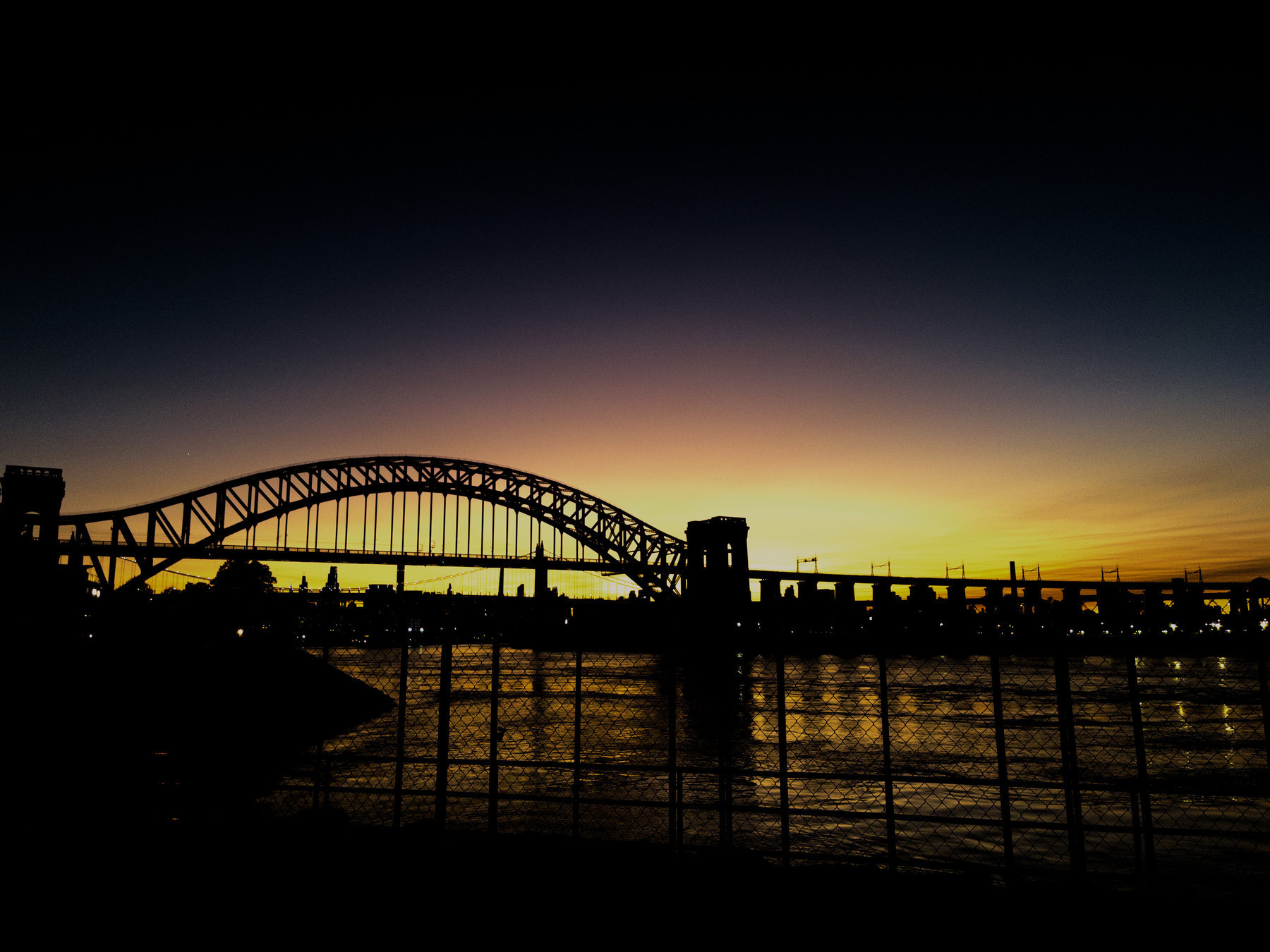 Auto-generated description: A silhouetted bridge spans across a body of water at dusk, with a vibrant sunset sky in the background.