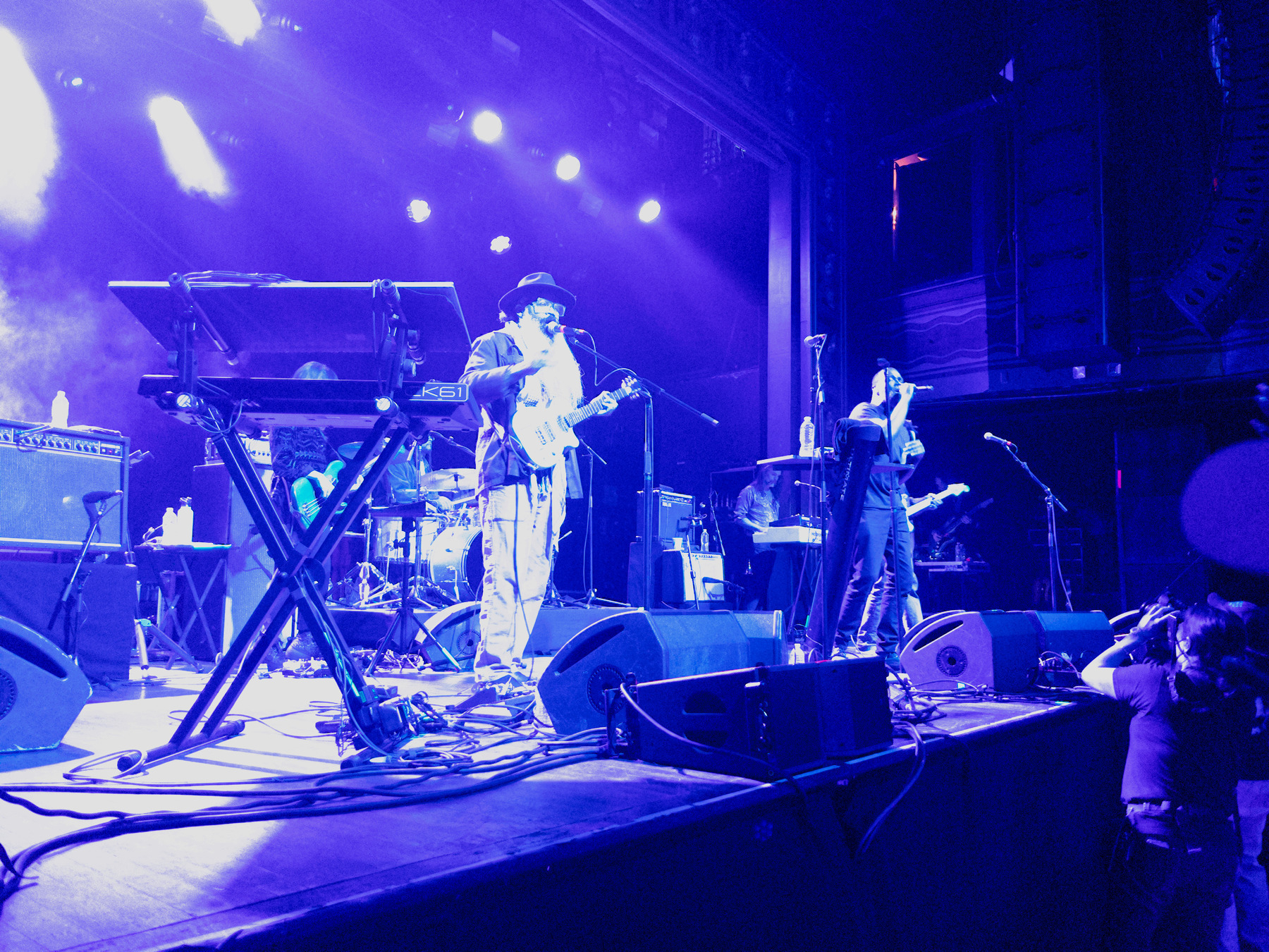 Auto-generated description: A band performs on stage under blue and purple lighting, featuring guitarists and a keyboardist.