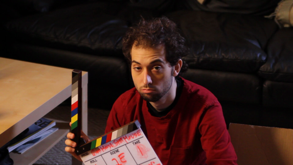 Me, a white man, years ago. I have messy hair, thin stubble, and a tired, sunken expression. I hold a film clapper. You can make out that it reads "Ghost house Shit house scene 2E take 1"