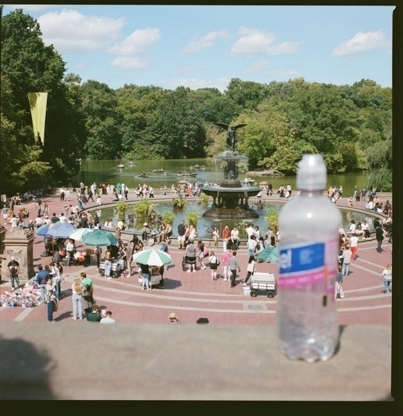 A park square with many dozens of people enjoying the day, shot from above. There's a fountain in the middle, and behind that is green water. People in row boast glide across the surface, and there's green trees behind on the opposite bank. In the extreme forground, out of focus, is the top of a low wall overlooking the square. A plastic water bottle rests atop it.