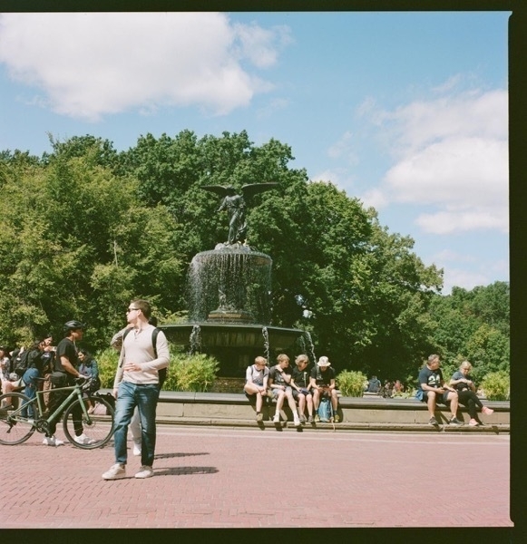 It's a bright day with a metal foundtain as the focus. The fountain has a decorative angle on top. There's red cobbles on the ground, and trees in the background. Several people of various ethnicities mill around and sit on the lip of the fountain. 