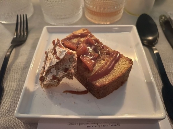 Close up of a thick, brown cake, almost zuccini bread looking, with a dark red jam with a fruit that looks like strawberries but isn't. There's a half eaten cookie with some of the jam on it and sugar coating. All on a white plate.