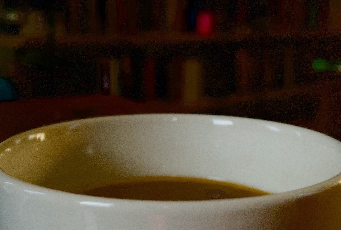 A close up, grainy picture of a white coffee mug in the bottom of the frame. You can just see the brown liquid inside. Beyond, and occupying most of the top of the frame, is a dark, out of focus bookshelf.