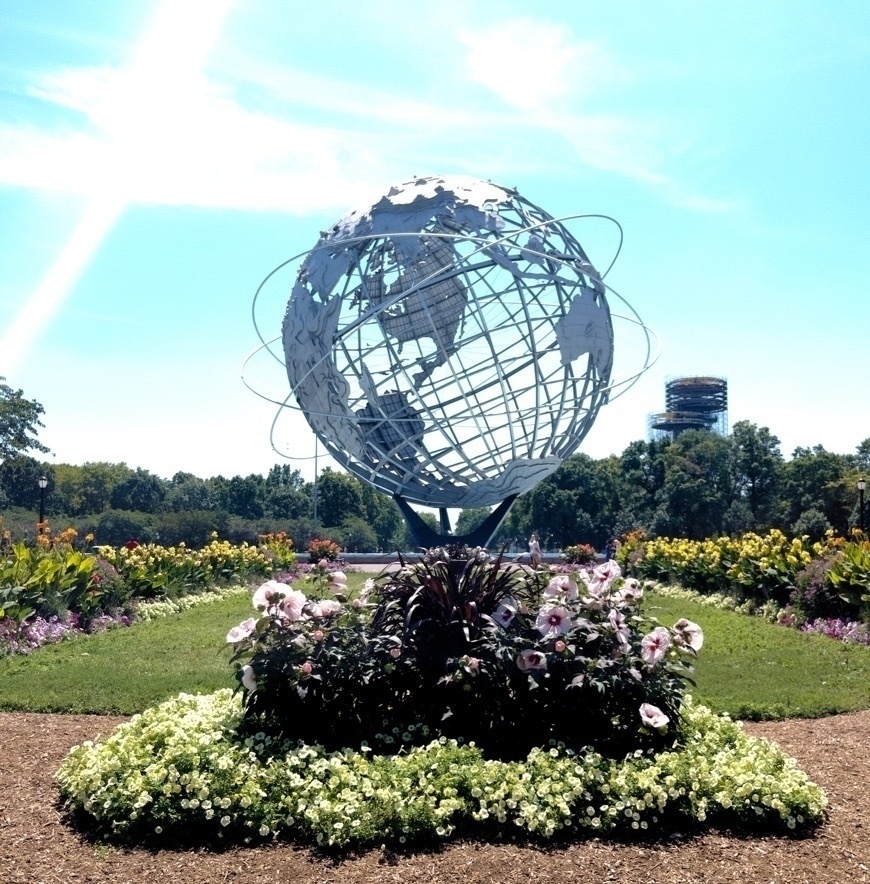 A large metal globe sculpture stands in a landscaped garden with flowers and greenery surrounding it.