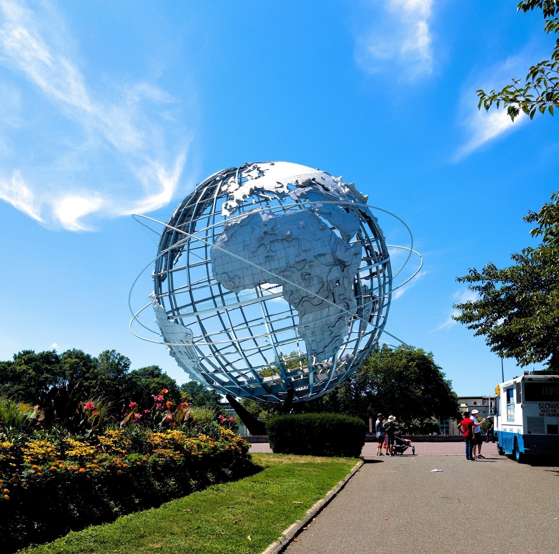 A large metallic globe sculpture stands in an outdoor park setting under a clear blue sky. Next to it, in the midground, is an ice cream truck, with a short line of people waiting outside.