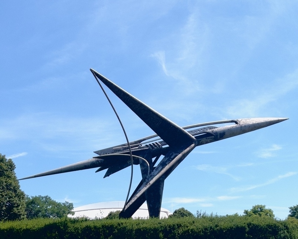 A sleek, futuristic rocket sculpture is displayed against a clear blue sky and greenery.