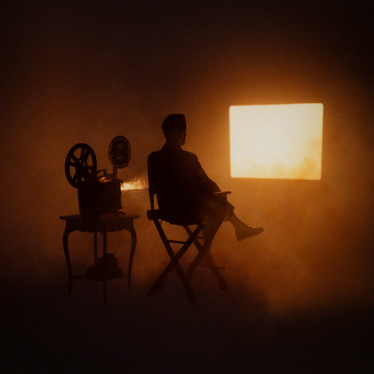 Auto-generated description: A person sits on a chair next to a film projector, facing a bright screen in a dark room.