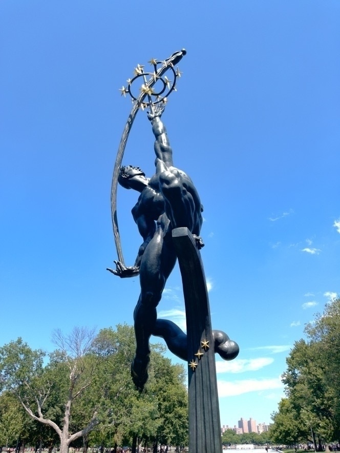 A statue of a muscular figure reaches skyward, holding a sphere with stars, set against a backdrop of trees and a clear blue sky.