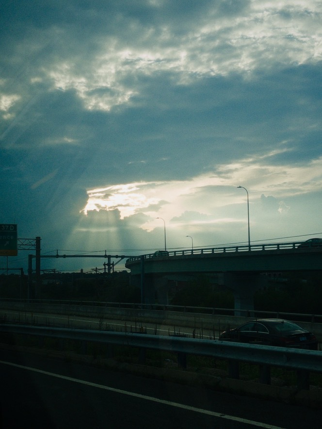 Auto-generated description: Sun rays break through thick clouds over an elevated highway with streetlights and cars.