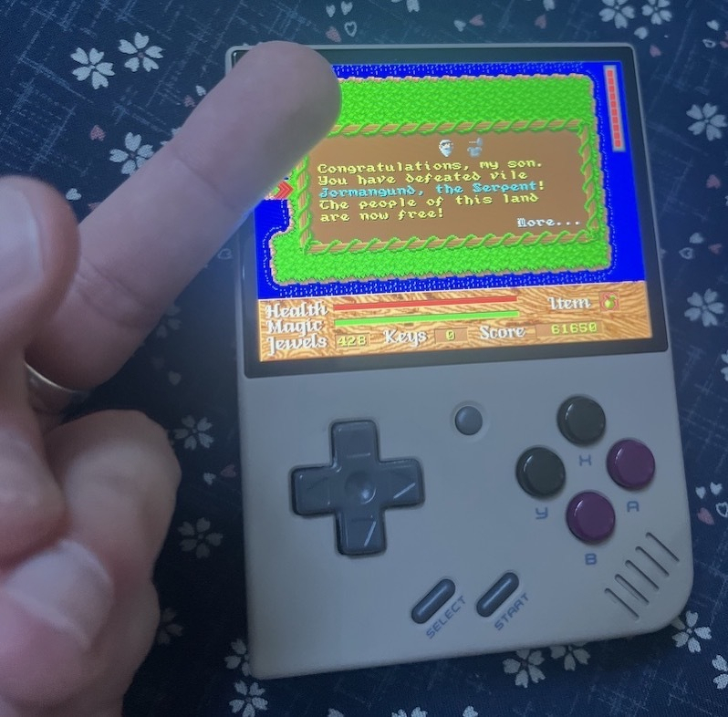 Auto-generated description: A handheld gaming device displays a victory screen from a game called The Serpent, while a hand shows a middle finger gesture in front of it.