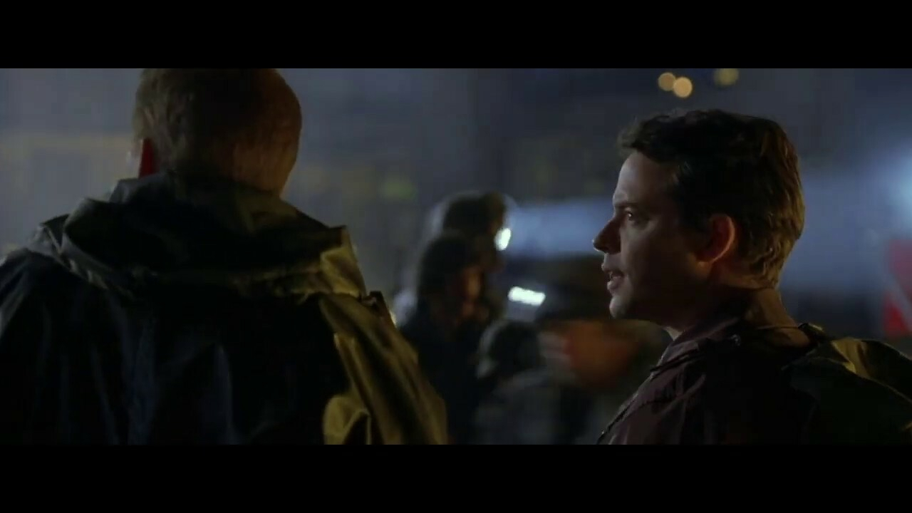 Auto-generated description: Two people in a dimly lit setting are engaged in conversation while others move in the background.