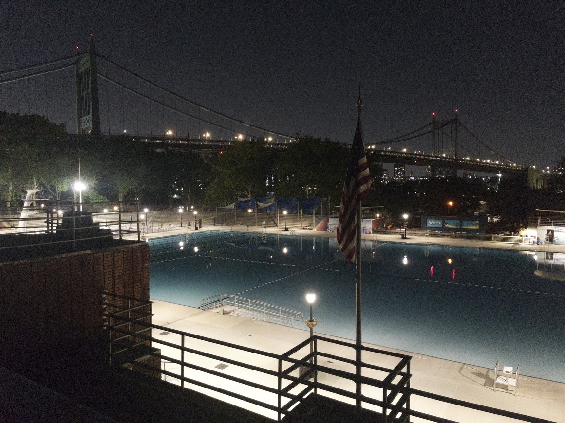 Auto-generated description: A nighttime scene features an illuminated outdoor swimming pool with an American flag and a suspension bridge in the background.