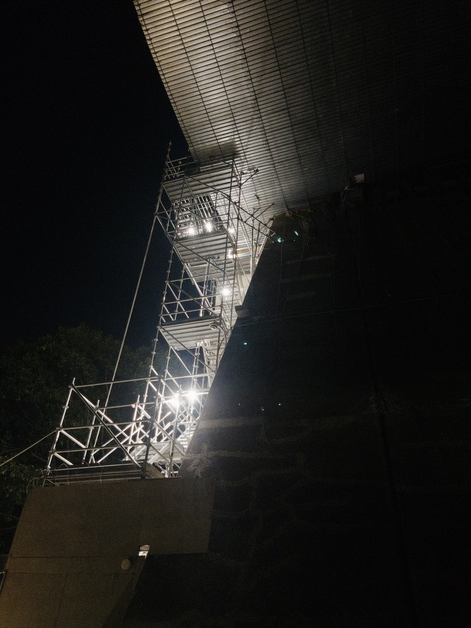 Auto-generated description: A metal scaffolding structure illuminated by bright lights is set up against a dark wall and under an overhead ceiling at night.