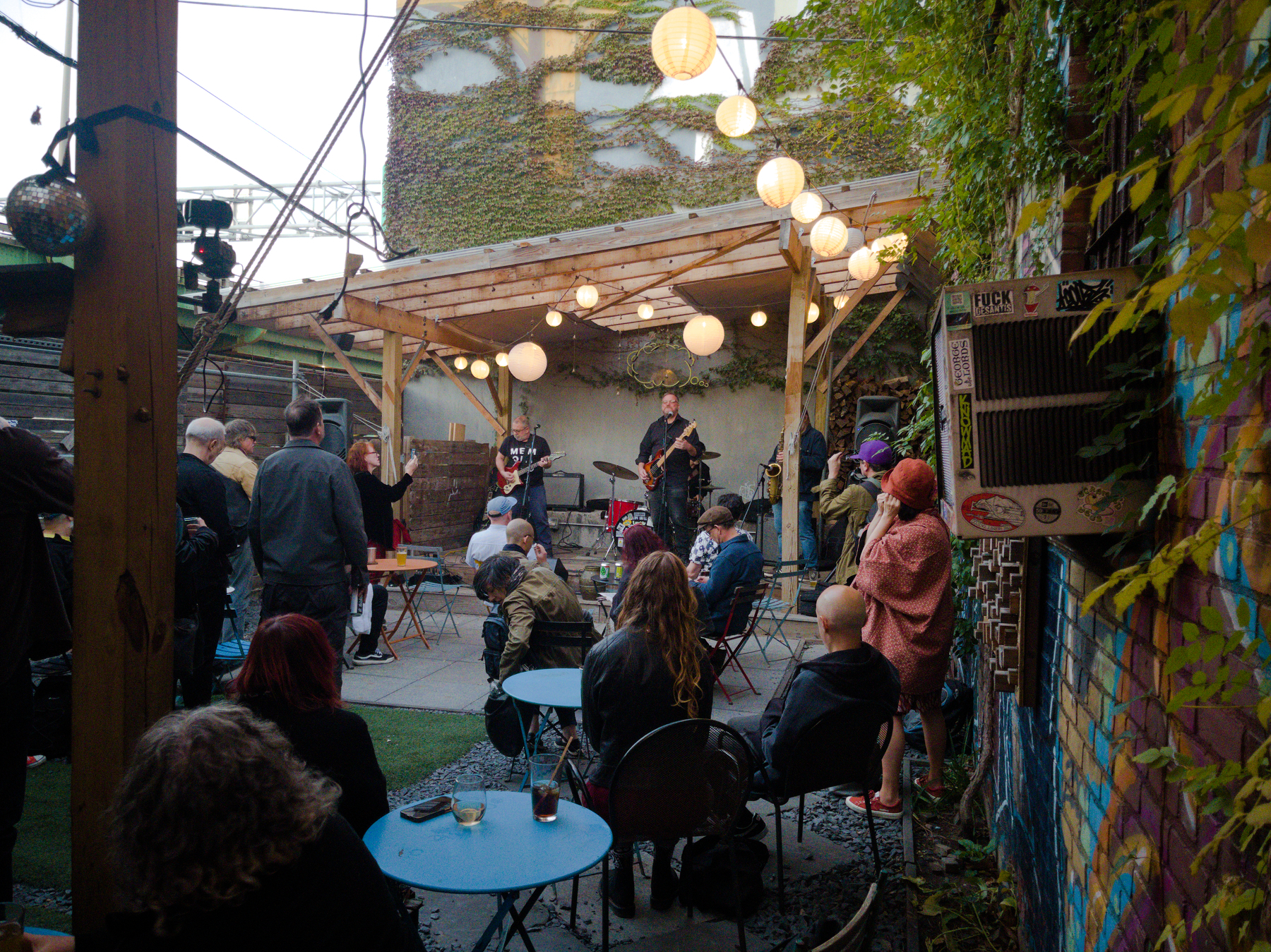 A small crowd is gathered in an outdoor courtyard with string lights and paper lanterns to watch a live band perform on a wooden stage.