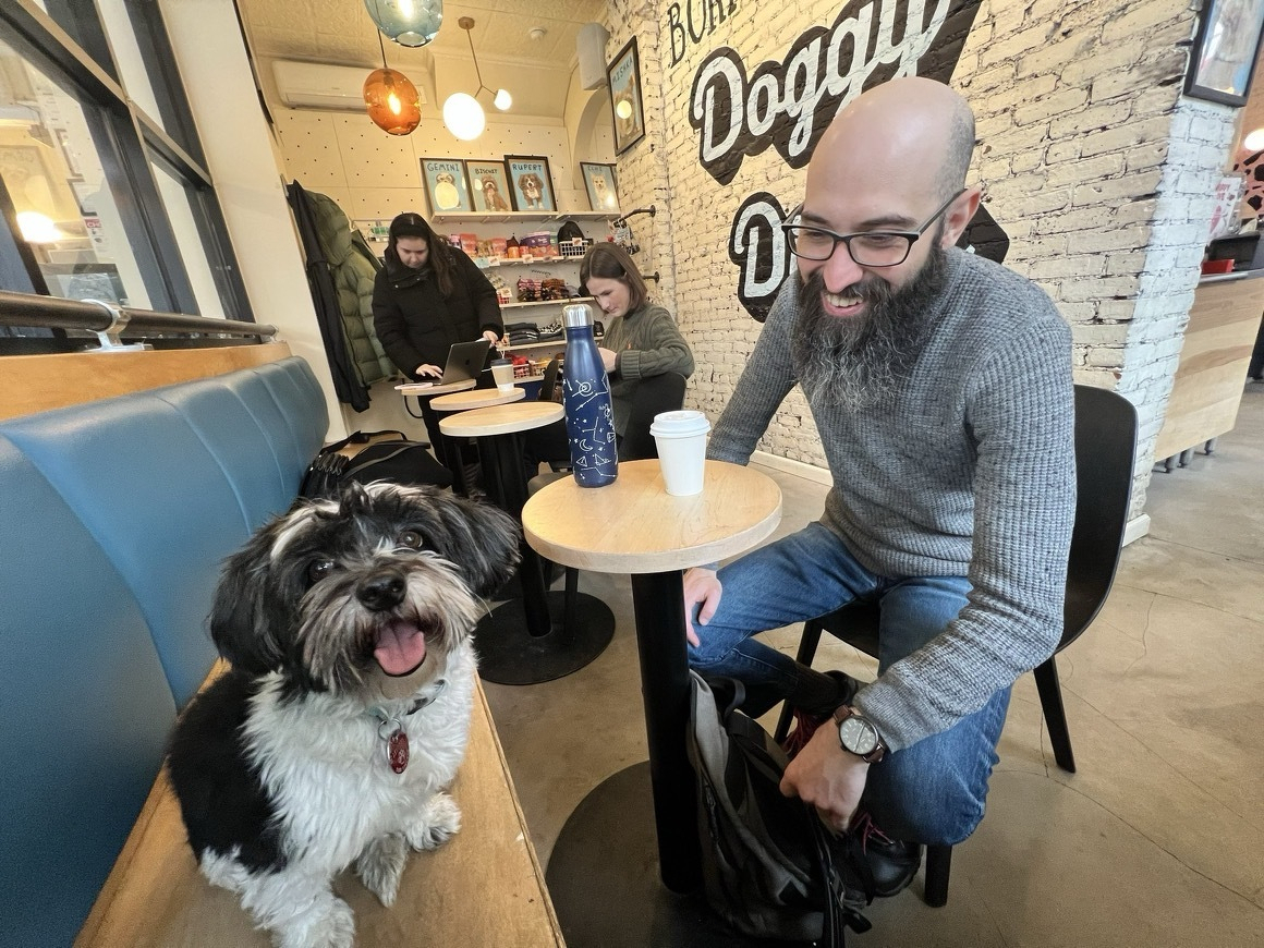 Auto-generated description: A man with a beard is sitting at a table in a cafe called Doggy with a dog sitting beside him on a bench.