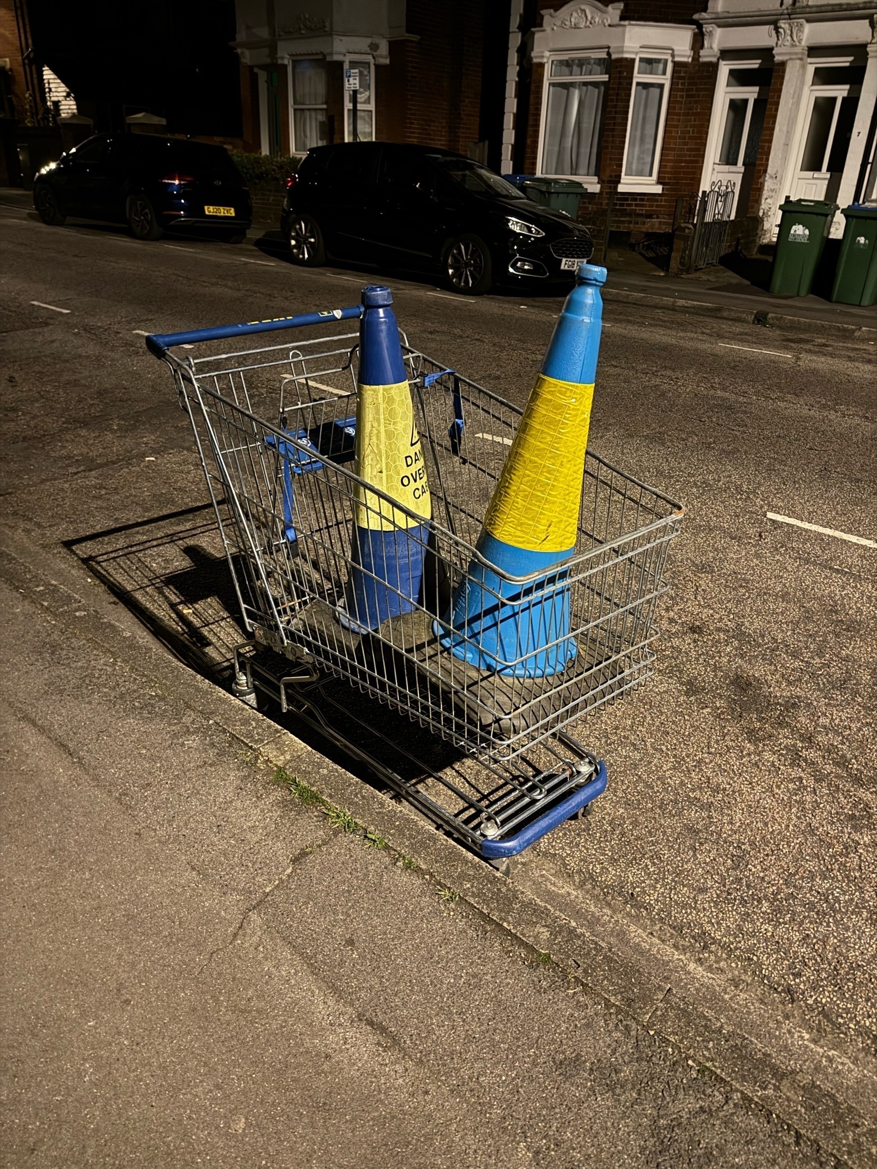 Supermarket trolley in the side of the road abandoned with two blue cones in the trolley with text warning about overhead cables likely dumped by local students  
