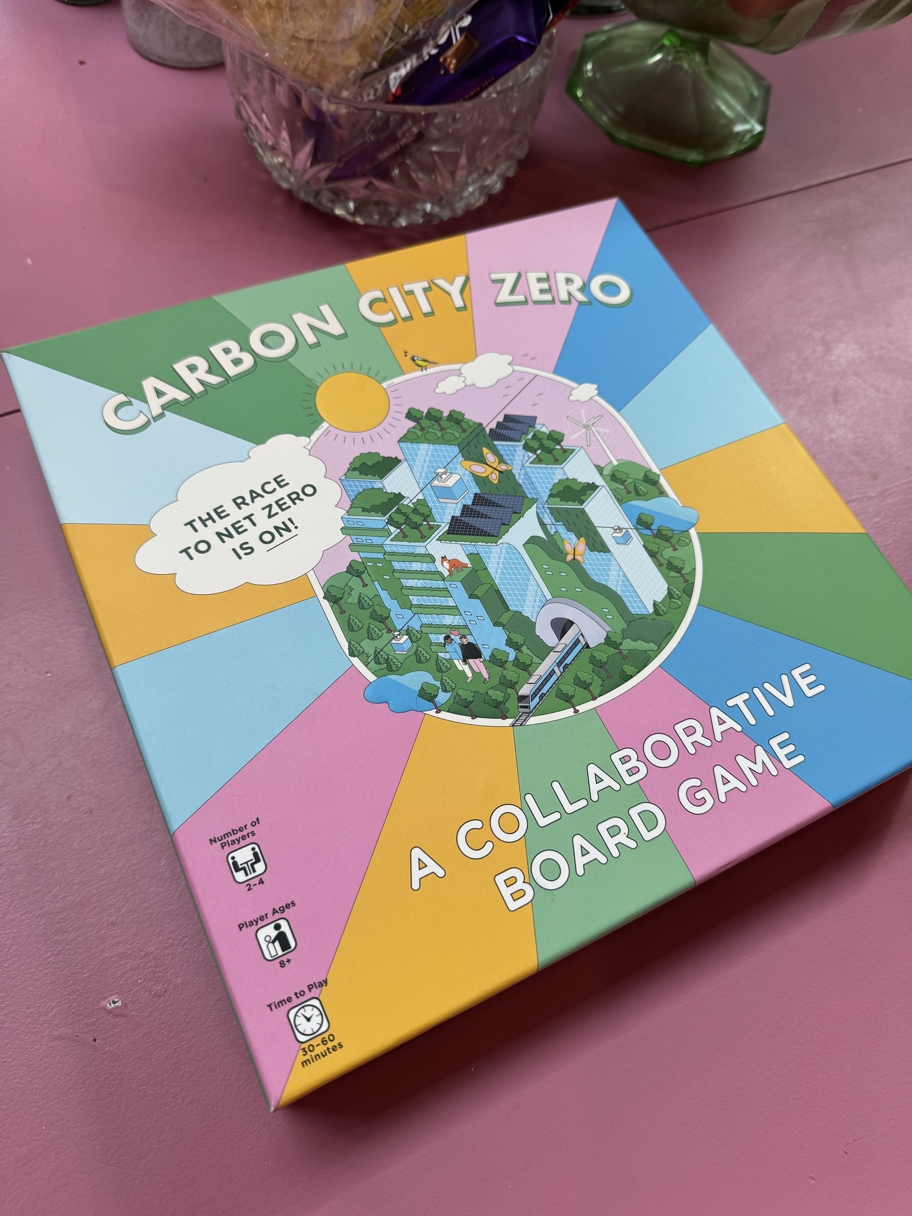 A colorful board game box titled "Carbon City Zero: A Collaborative Board Game" is displayed on a pink surface.
