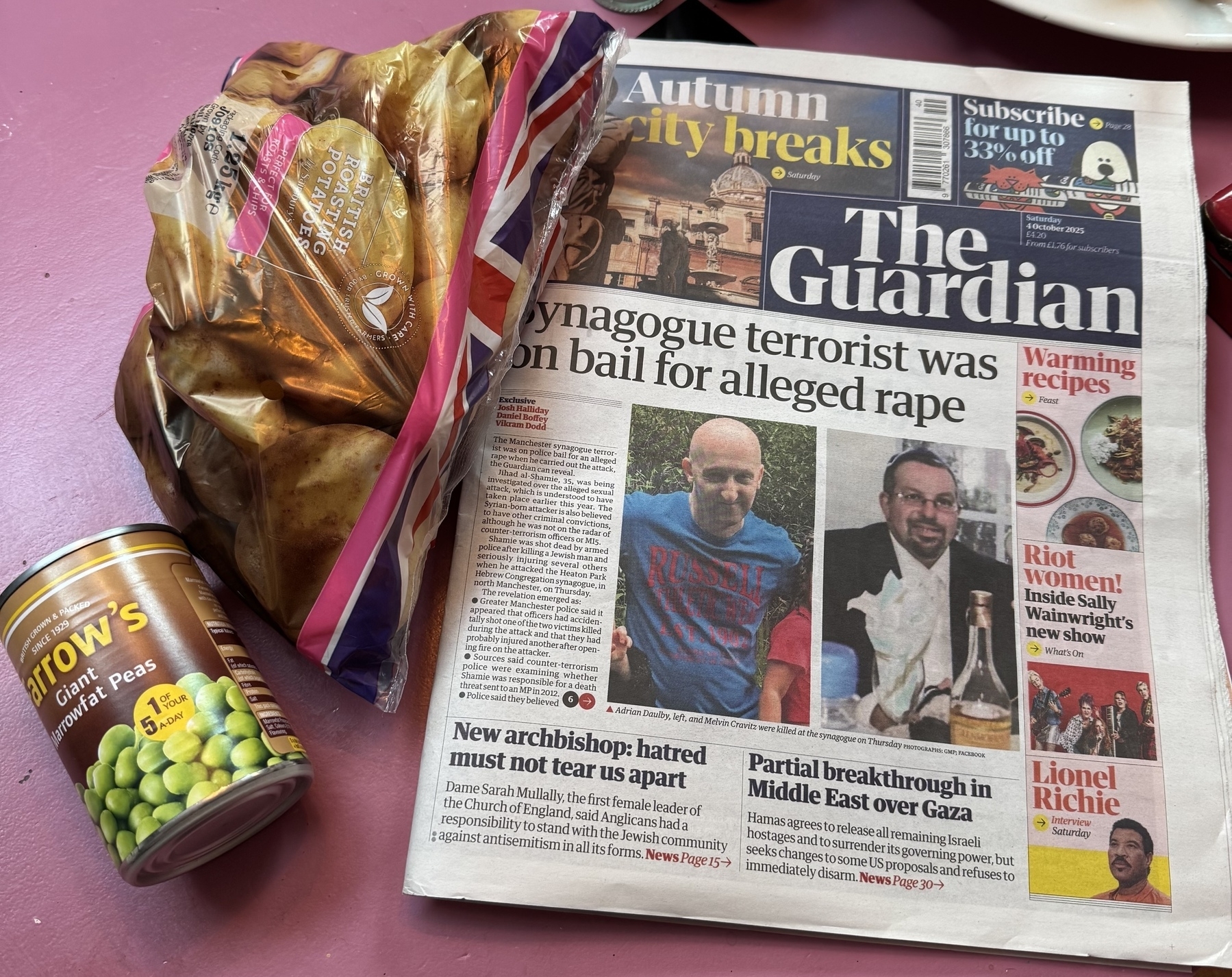 A newspaper, a can of peas, and a bag of potatoes are arranged on a pink surface.