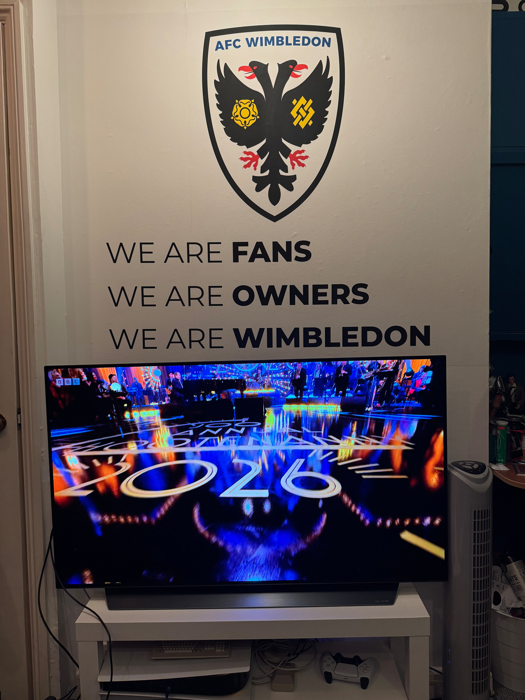 A television screen displays vibrant, colorful graphics in front of a wall with the AFC Wimbledon logo and motivational text about fans and ownership.