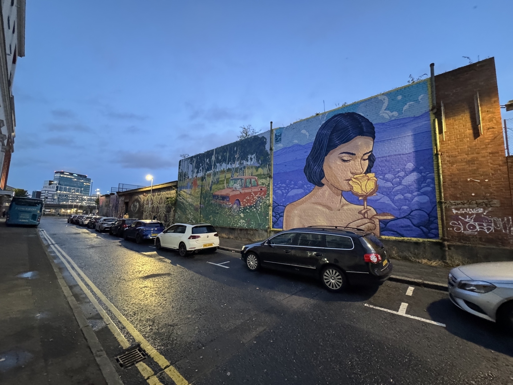 A city street features parked cars and a large mural of a woman smelling a yellow rose on the side of a building.
