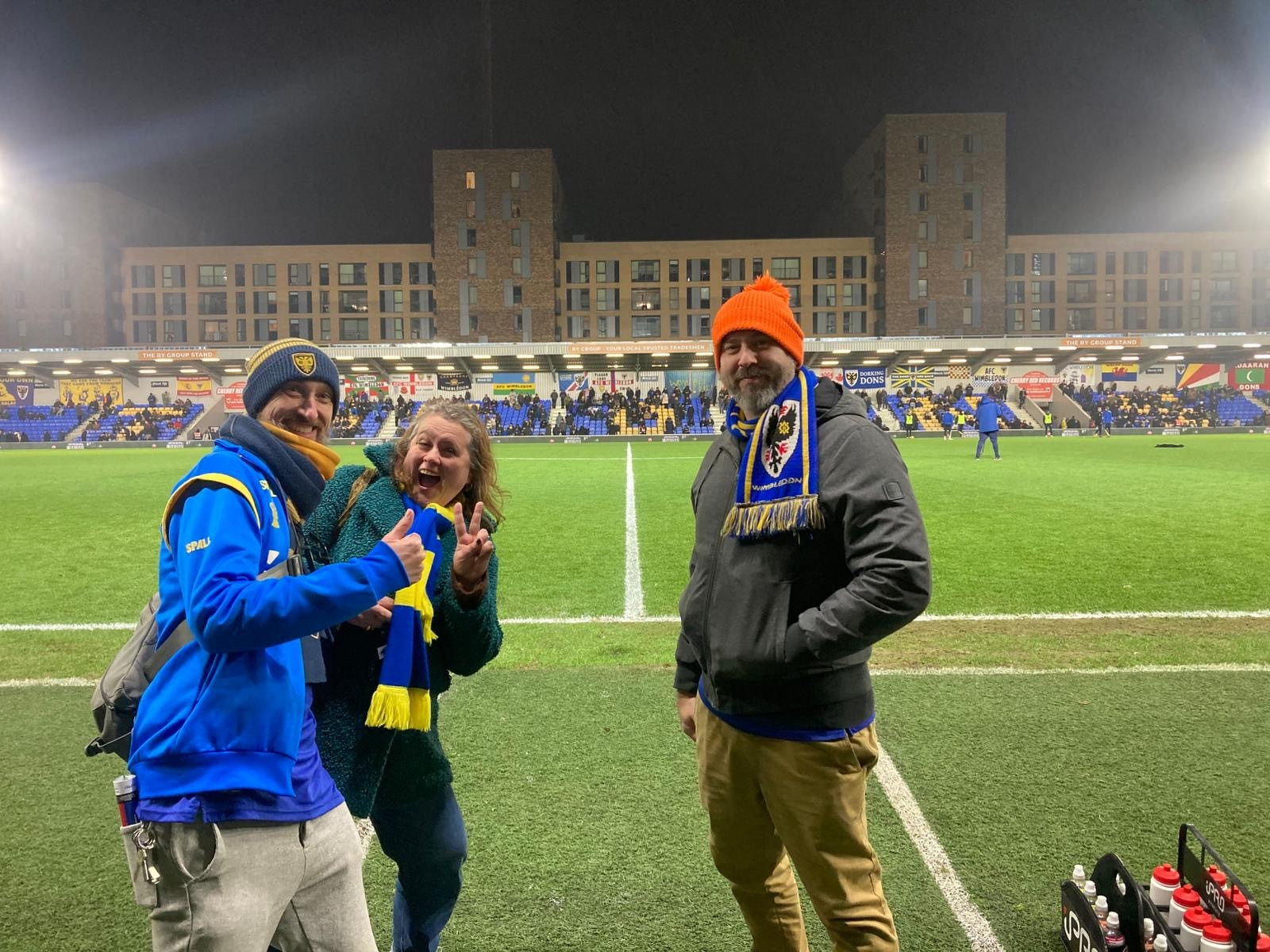 Three people are posing for a photo at a football stadium, dressed warmly with scarves and hats, with the field and stands visible in the background.