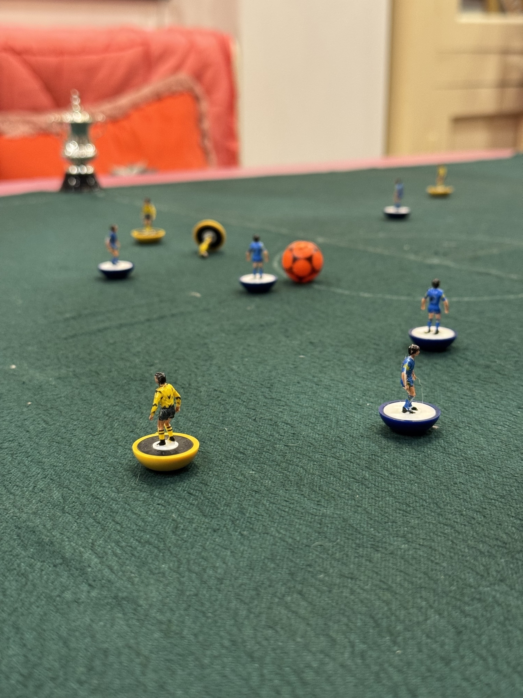 A tabletop soccer game setup features miniature players on a green field with a small orange ball.