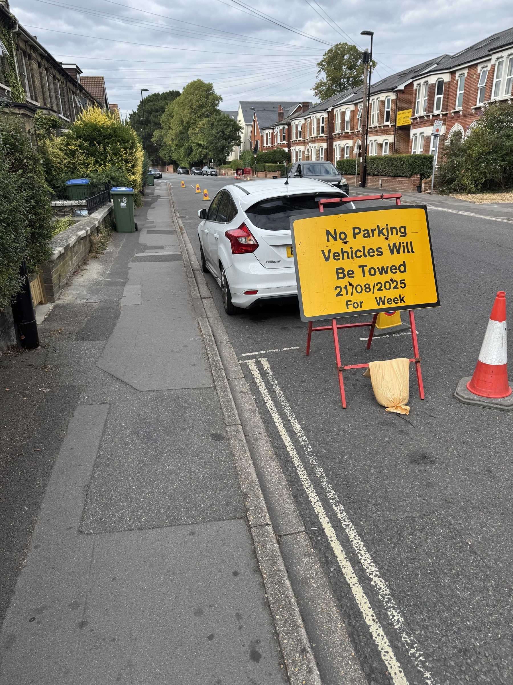 A parked white car is next to a "No Parking" sign and traffic cones on a suburban street.