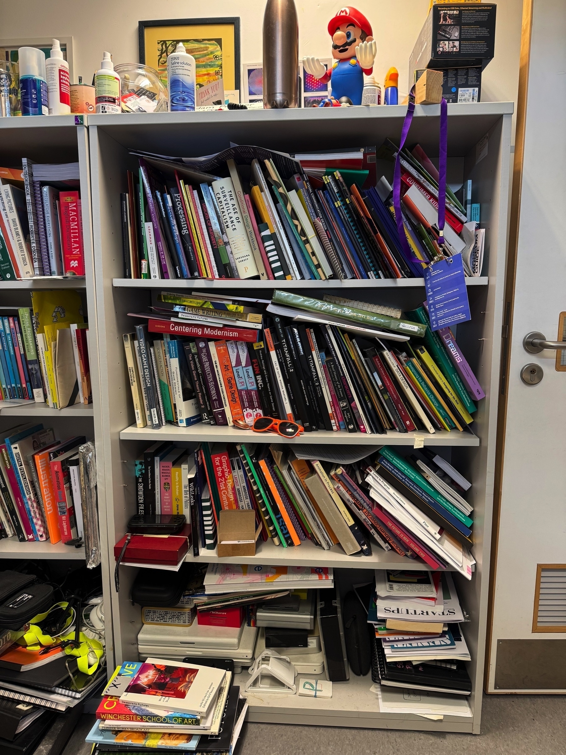 A cluttered bookshelf overflowing with an array of books, folders, and miscellaneous items, including a figure of a popular video game character on top.