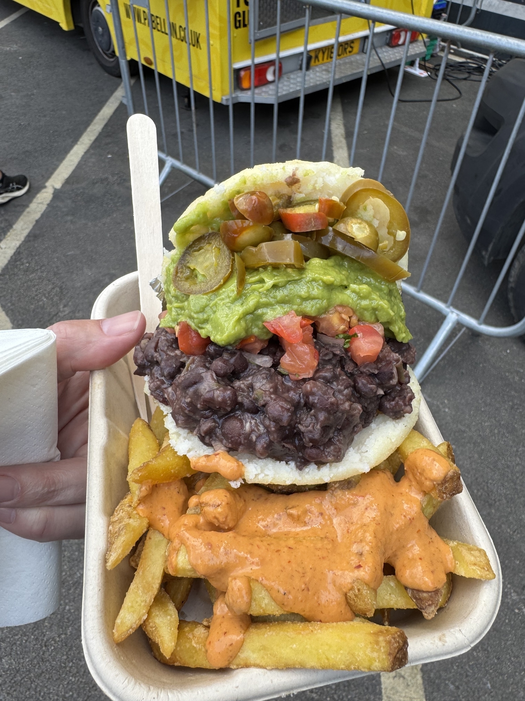A hand holds a tray containing loaded fries topped with sauce and a sandwich filled with black beans, guacamole, diced tomatoes, and jalapeños.