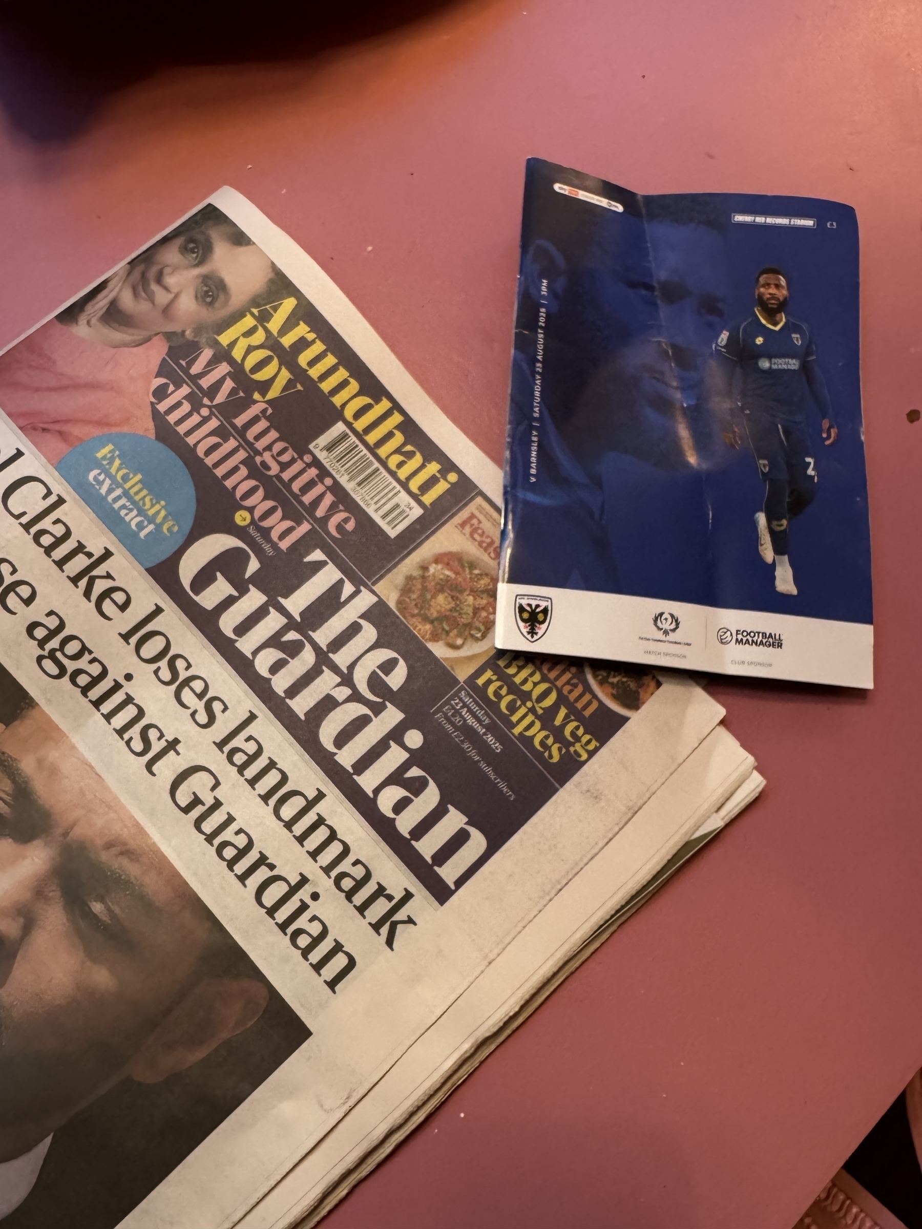 A Guardian newspaper and a football magazine featuring a man in a blue kit are placed on a pink surface.