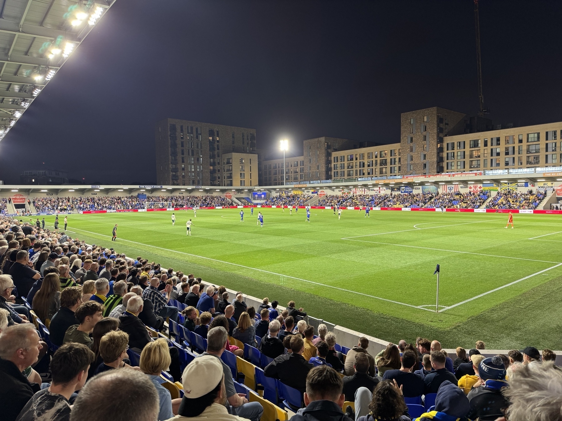 Fans watch a nighttime football match at Plough Lane (Cherry Red Records Stadium) with residential buildings in the background.
