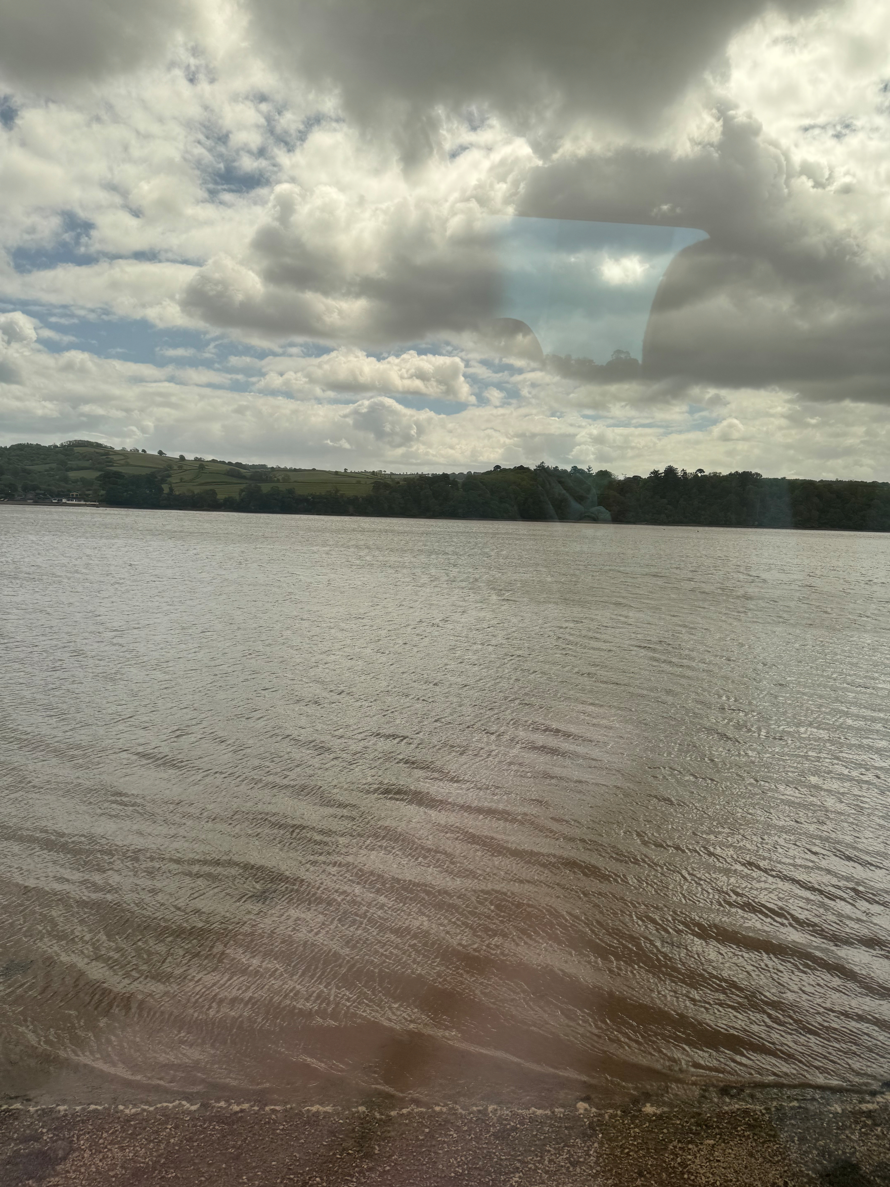A calm body of water stretches out under a cloudy sky, with a distant shoreline and rolling hills in the background.