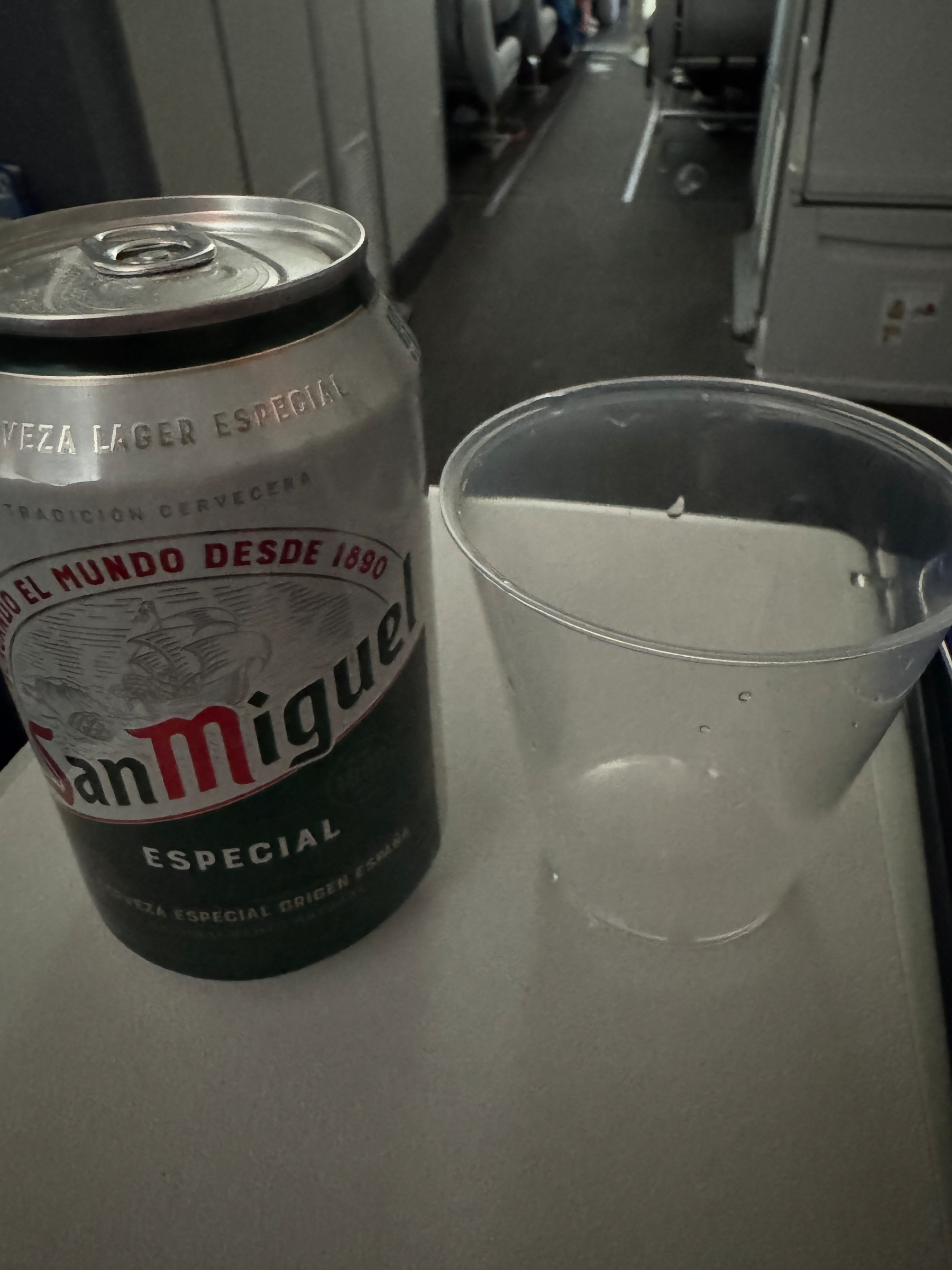 A can of San Miguel Especial beer and a clear plastic cup are placed on a tray table inside an airplane.