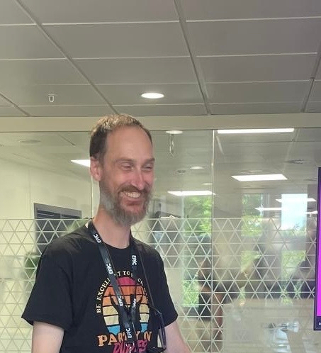 A man with a beard wearing a black t-shirt and a lanyard is smiling inside a modern office space with glass partitions.