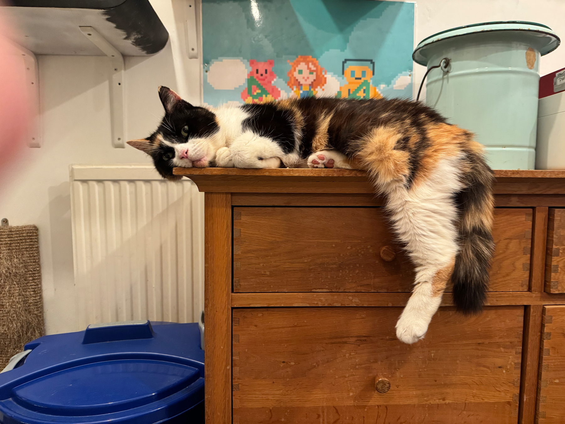 A calico cat is lounging on top of a wooden dresser with one leg hanging off the edge.