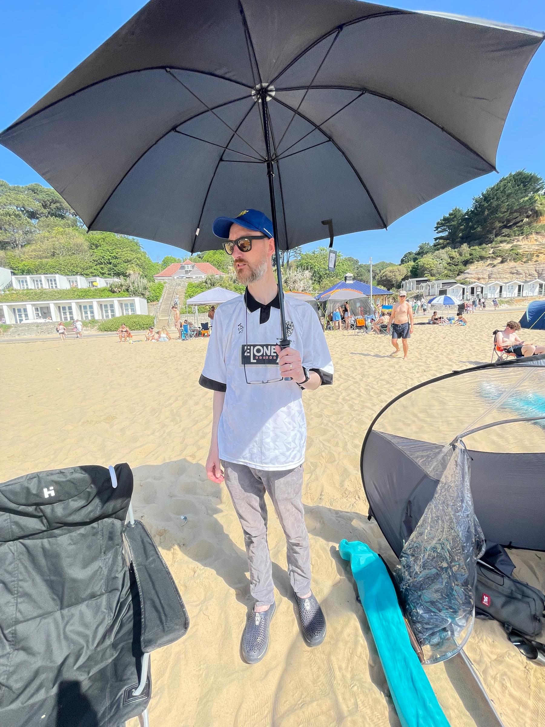 A person stands on a sandy beach holding a large umbrella, dressed casually with a blue cap and sunglasses, surrounded by beach chairs and people.