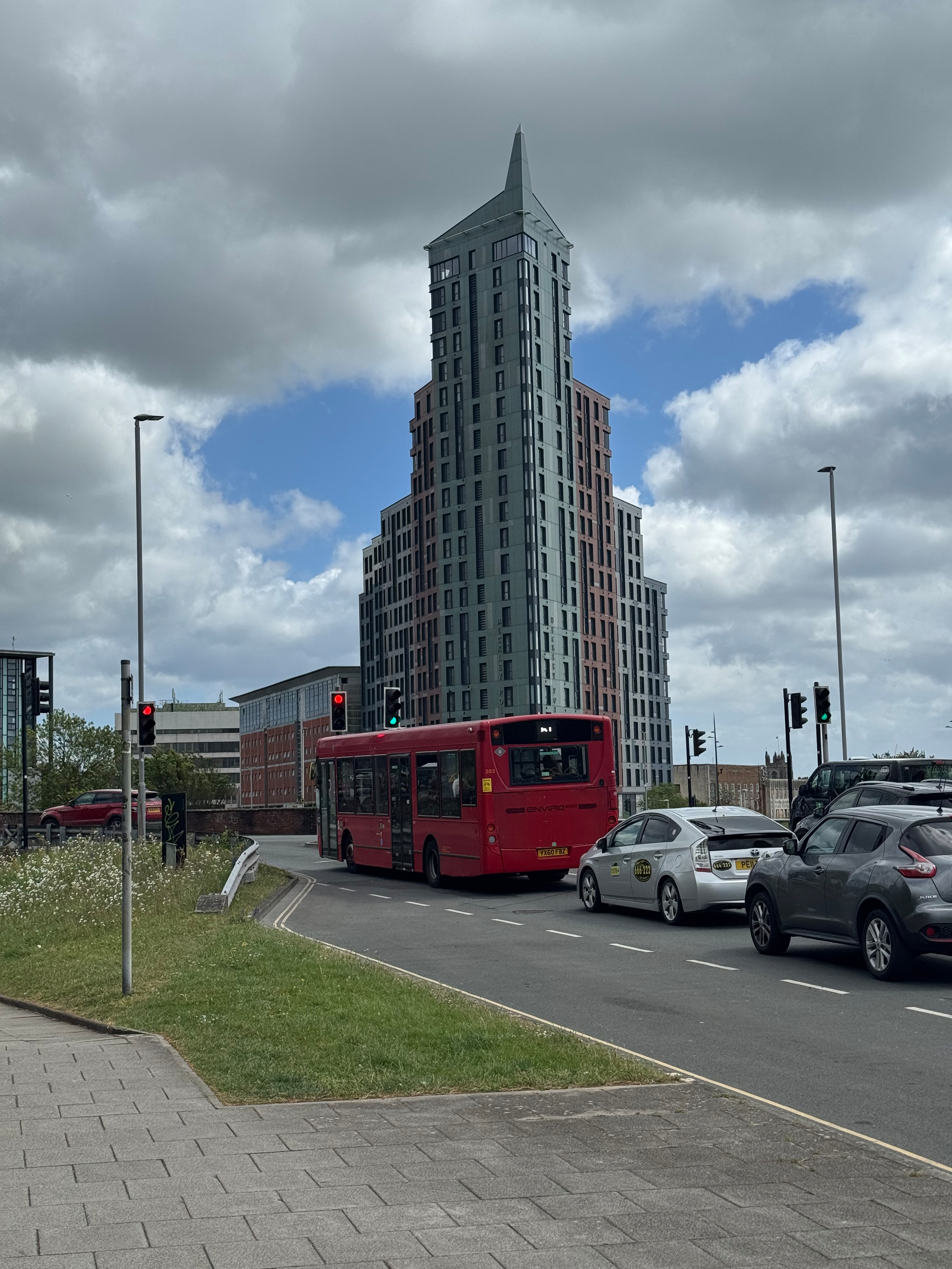 A red bus and several cars are stopped at a traffic light near a modern high-rise building on a cloudy day.