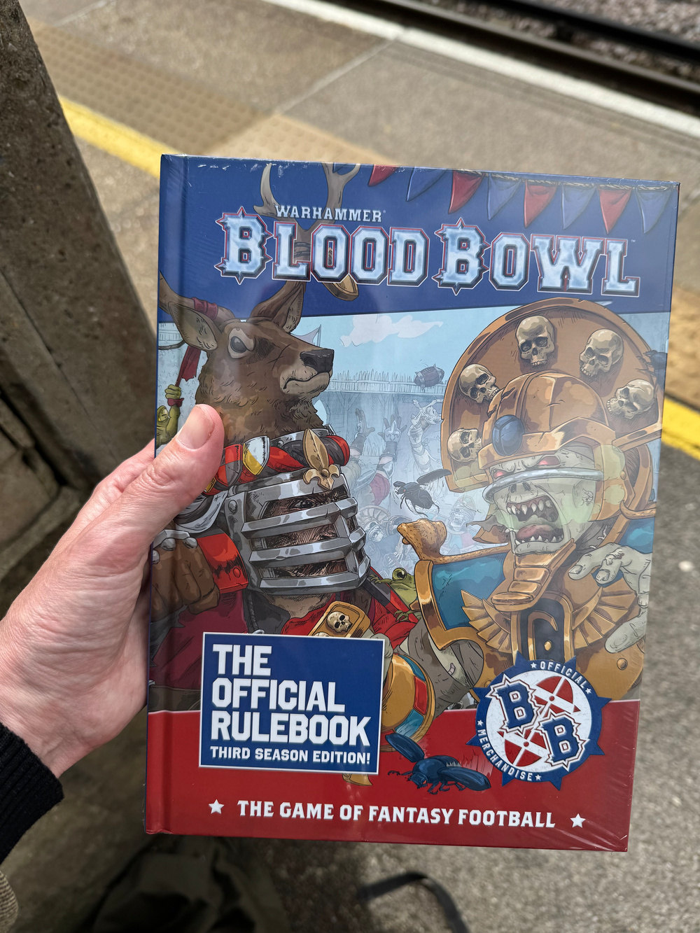 A person is holding the official rulebook for Warhammer's Blood Bowl, a fantasy football game, at a train station.