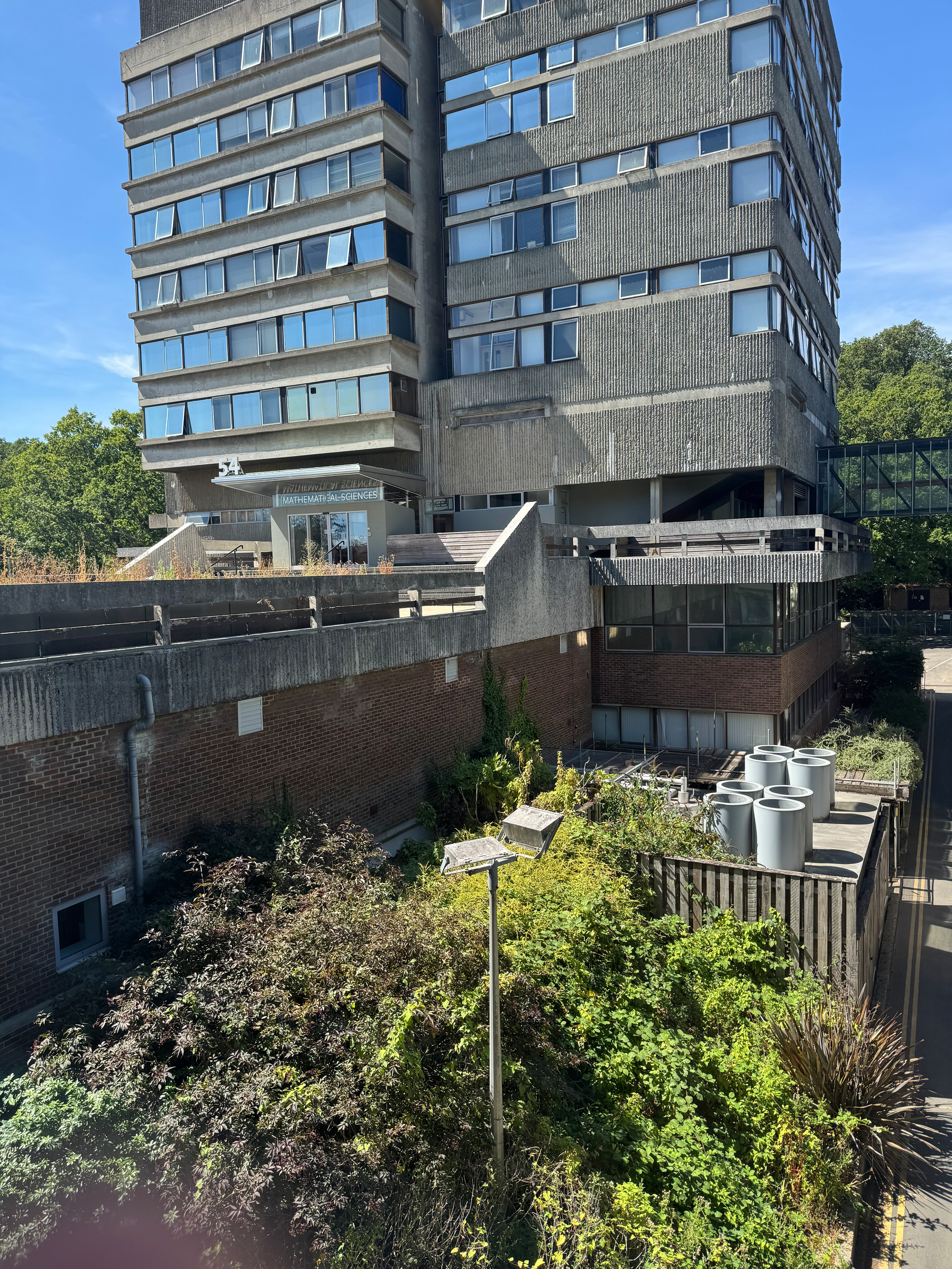 A multi-story building with a concrete and brick exterior is surrounded by greenery and outdoor utility structures under a clear blue sky.