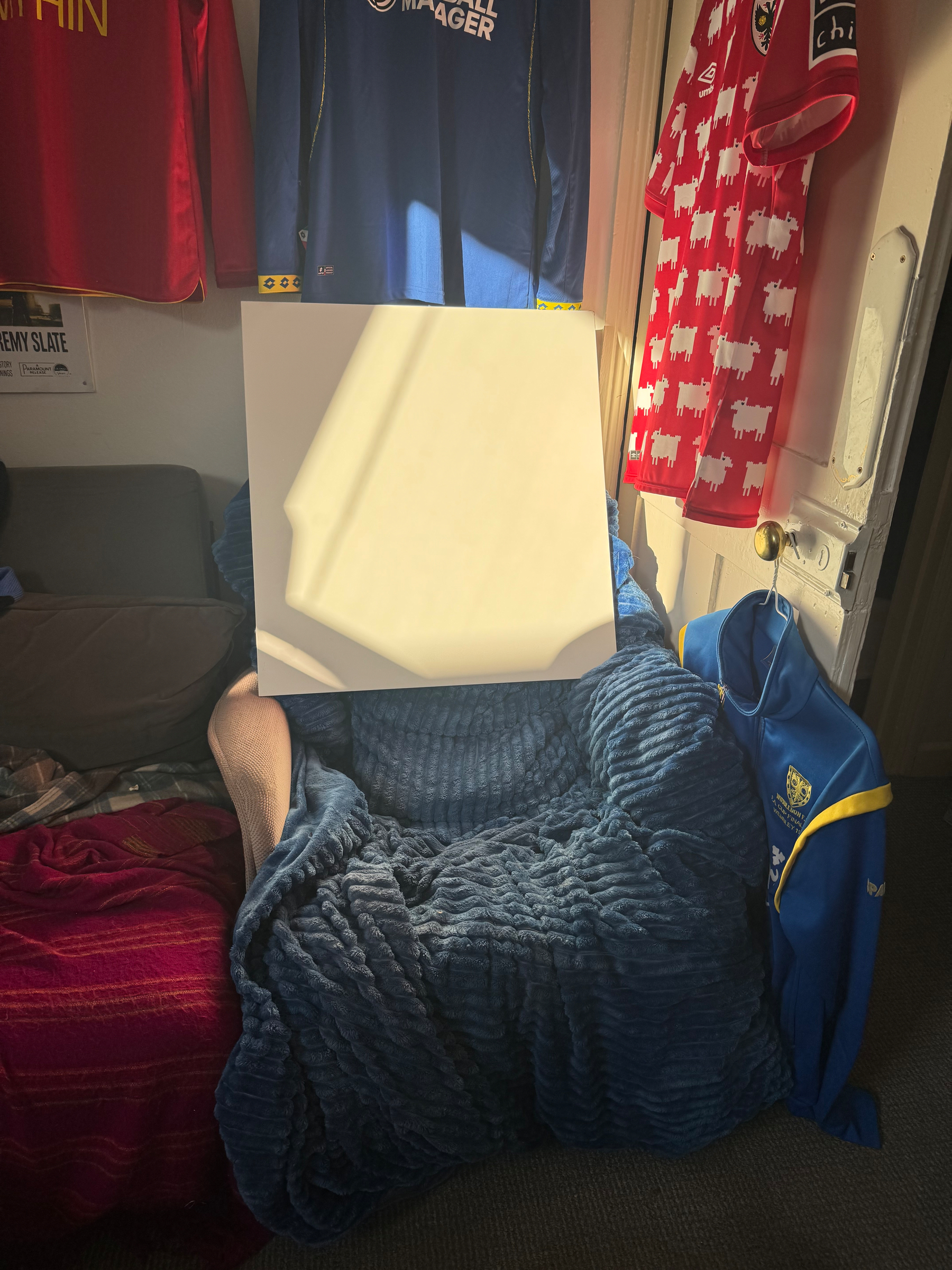 a cozy armchair draped with a soft blue blanket, with a white square placed in the sun surrounded by various colorful AFC Wimbledon shirts hanging on the walls.