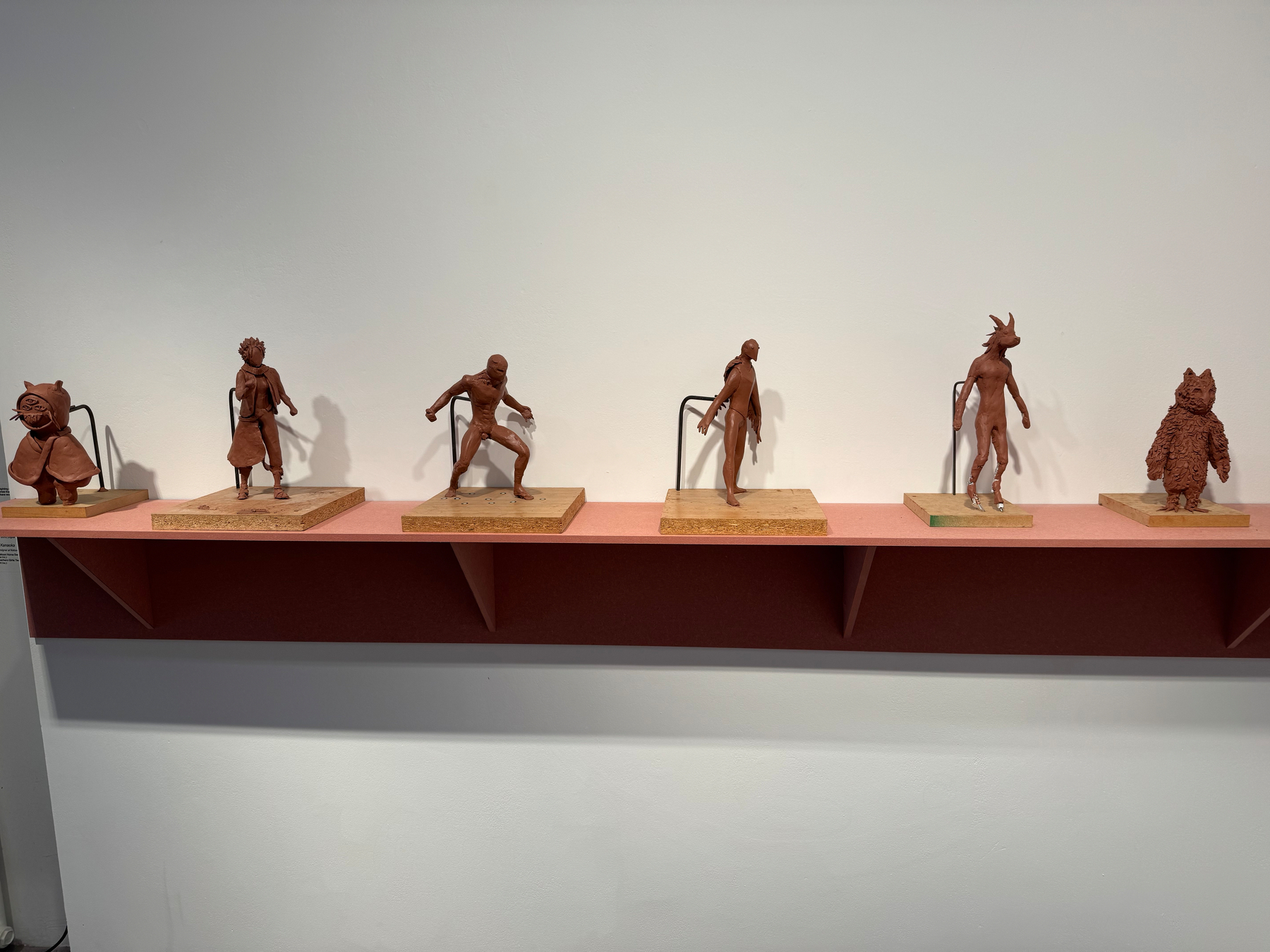 Six small, uniquely shaped clay sculptures are displayed on a shelf against a white wall.